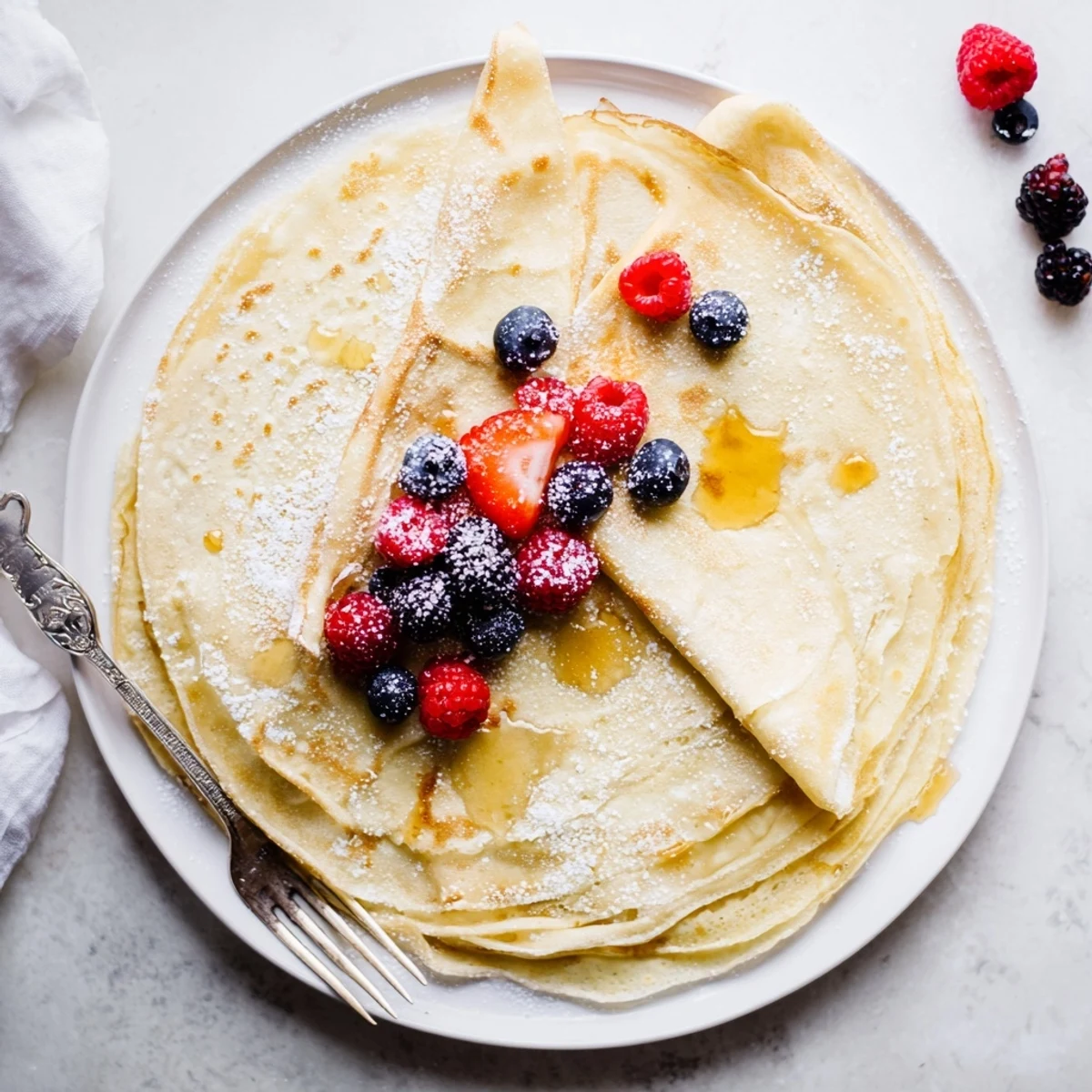 Stack of warm crepes with honey drizzle garnished with fresh berries and dusted powdered sugar